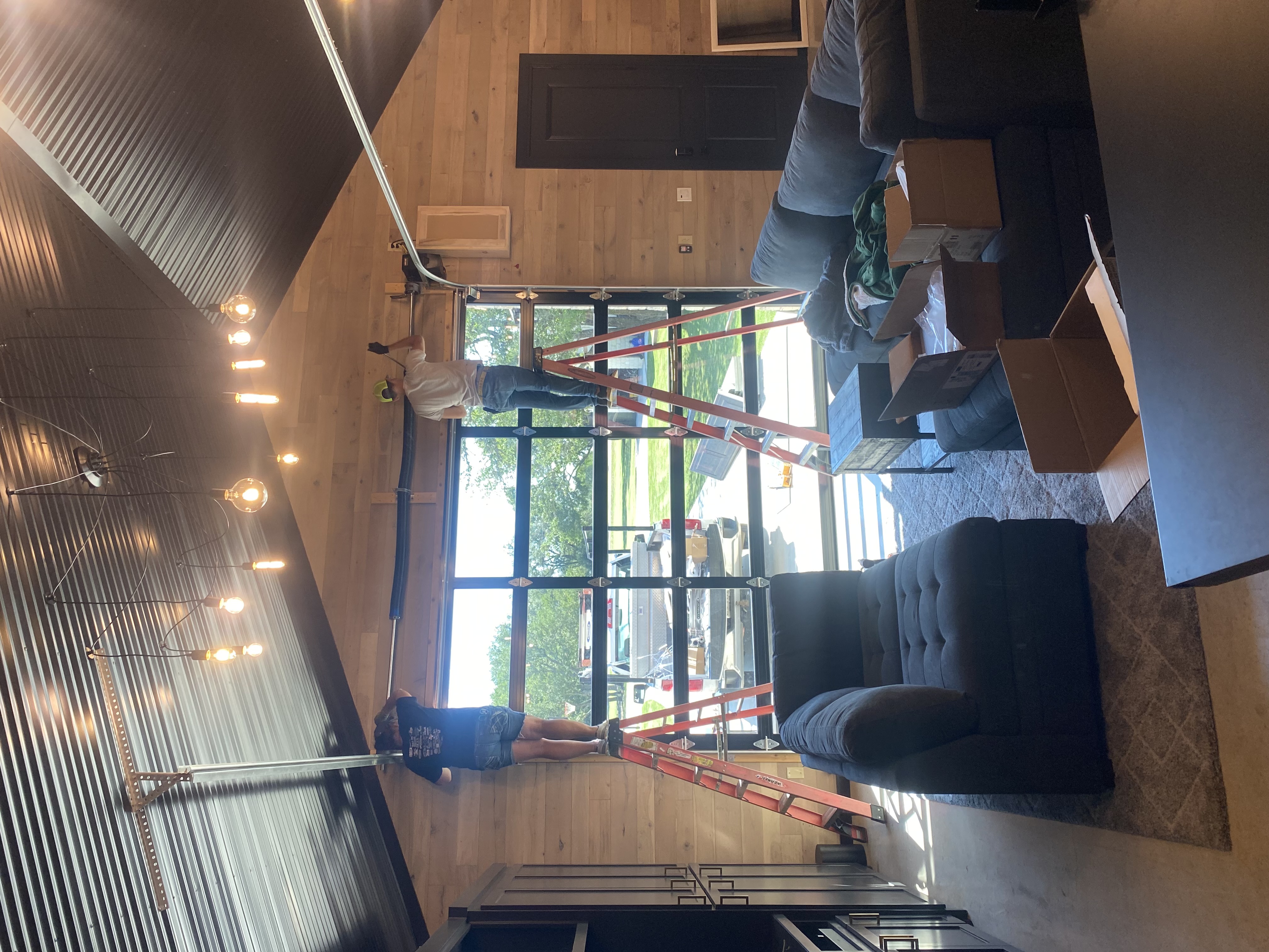 Two Palousek Overhead Doors technicians on ladders actively installing a full-view glass door inside a barndominium with corrugated metal ceiling and string lights